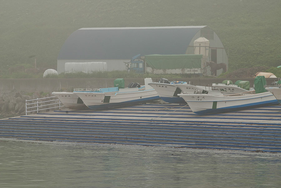 Moderne Fischerboote mit großen Motoren zeugen von der schweren Arbeit. Im Hintergrund eine Lagerhalle. Nebel liegt zwischen den Hügeln.